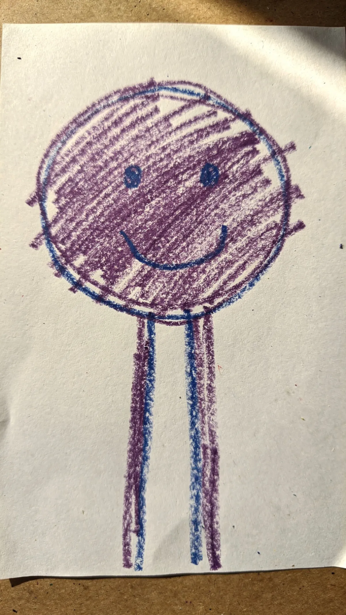 A child's purple and blue crayon drawing of a circle-head figure with two stick legs on white paper, typical of a 3-year-old's artwork