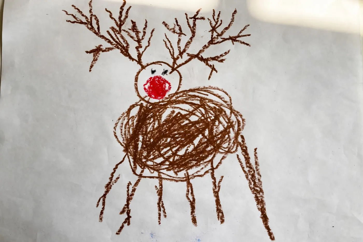 A child's crayon drawing on white paper of a brown reindeer with a scribbled oval body, a bright red nose, huge branching antlers and six legs of uneven lengths