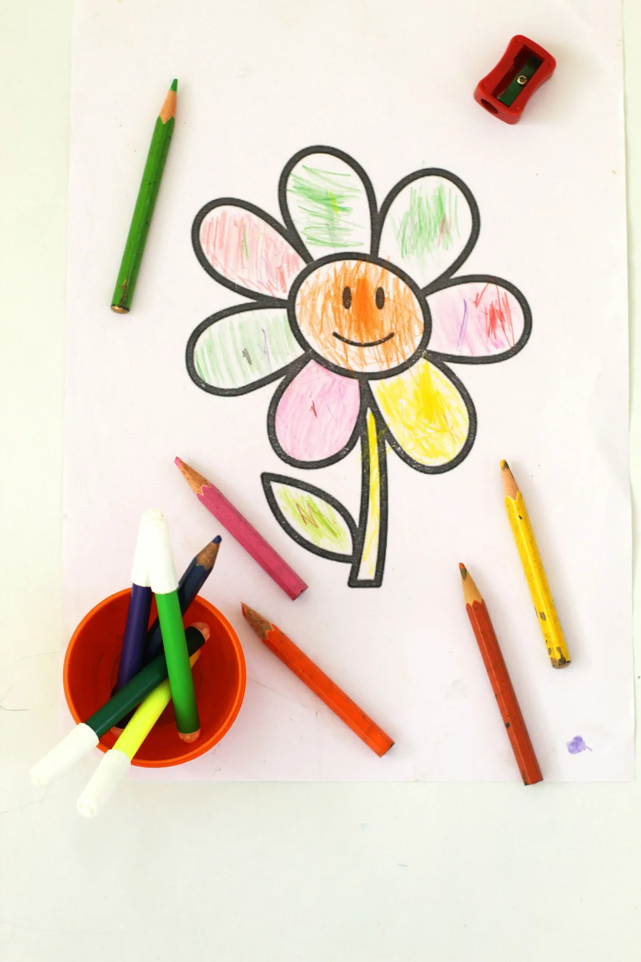 A child's colorful flower drawing on white paper with a smiley face center, colored pencils and a pencil sharpener scattered around it, overhead flat lay