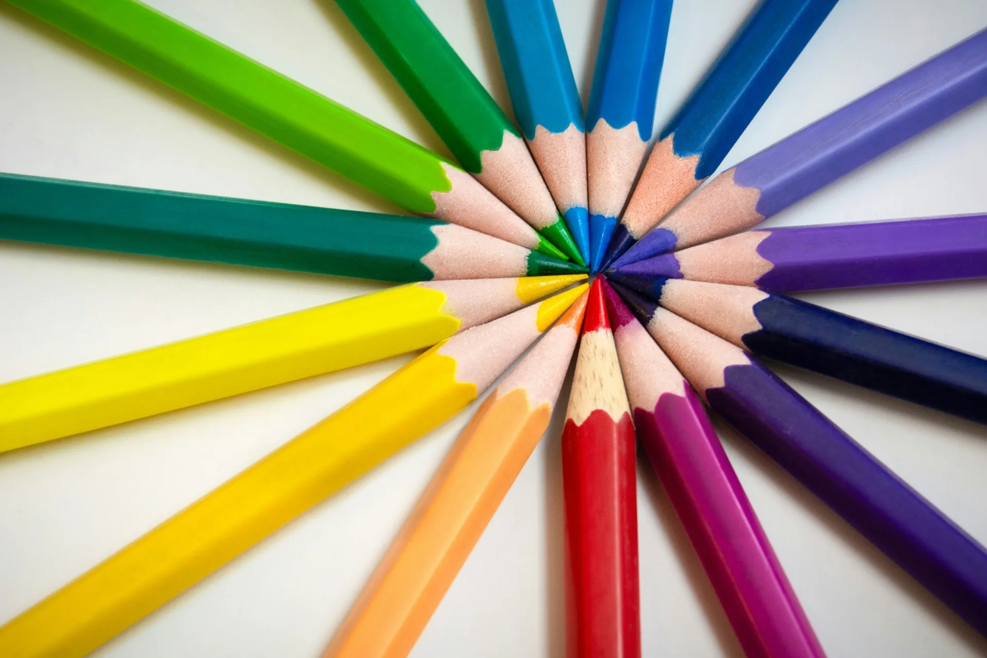 Colored pencils arranged in a sunburst fan pattern on a white surface, every color radiating from the center — green, blue, purple, red, orange, yellow