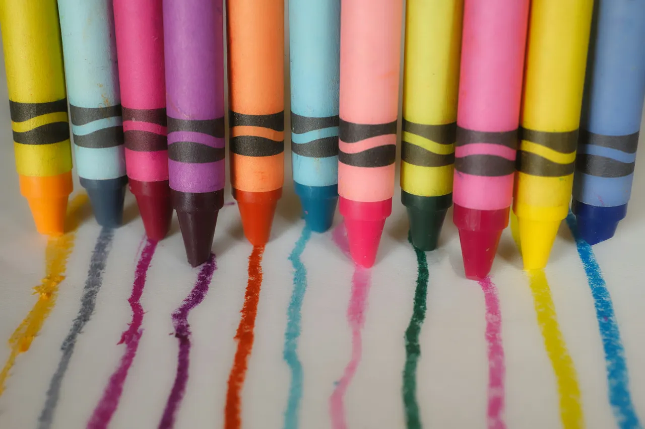 A row of colored crayons each drawing a line of its own color on white paper