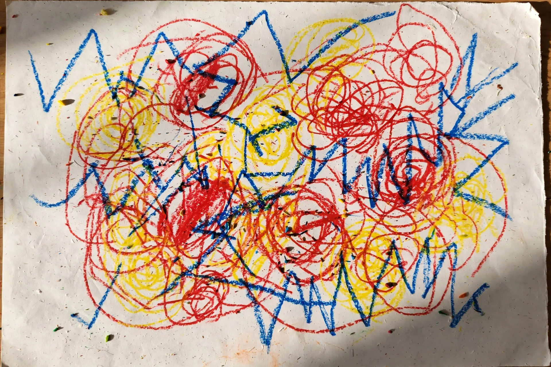 A toddler's overlapping red, yellow, and blue crayon scribbles on white paper — loops and zig-zags with no recognizable shapes