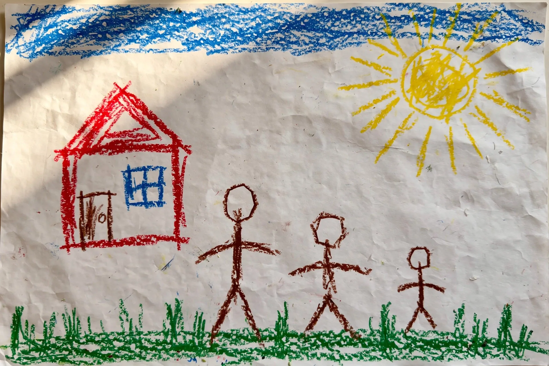 A child's crayon scene with a blue sky strip, a yellow sun with rays, a red house, and three stick-figure people of different heights on a green grass line