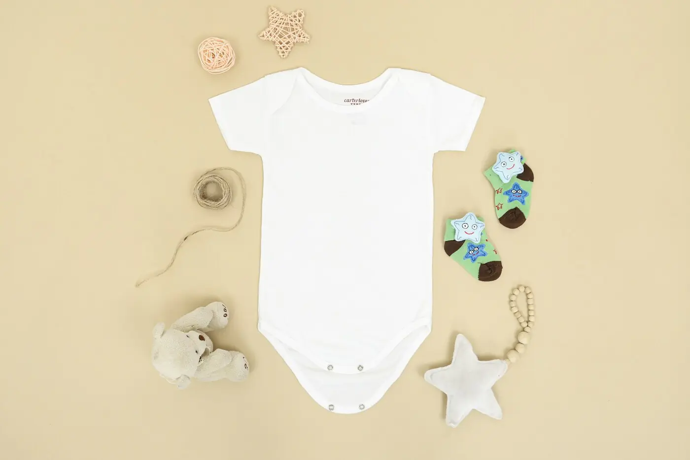 Overhead flat lay of a plain white newborn onesie on a soft beige background, surrounded by small baby socks, a knitted star and a tiny stuffed bear