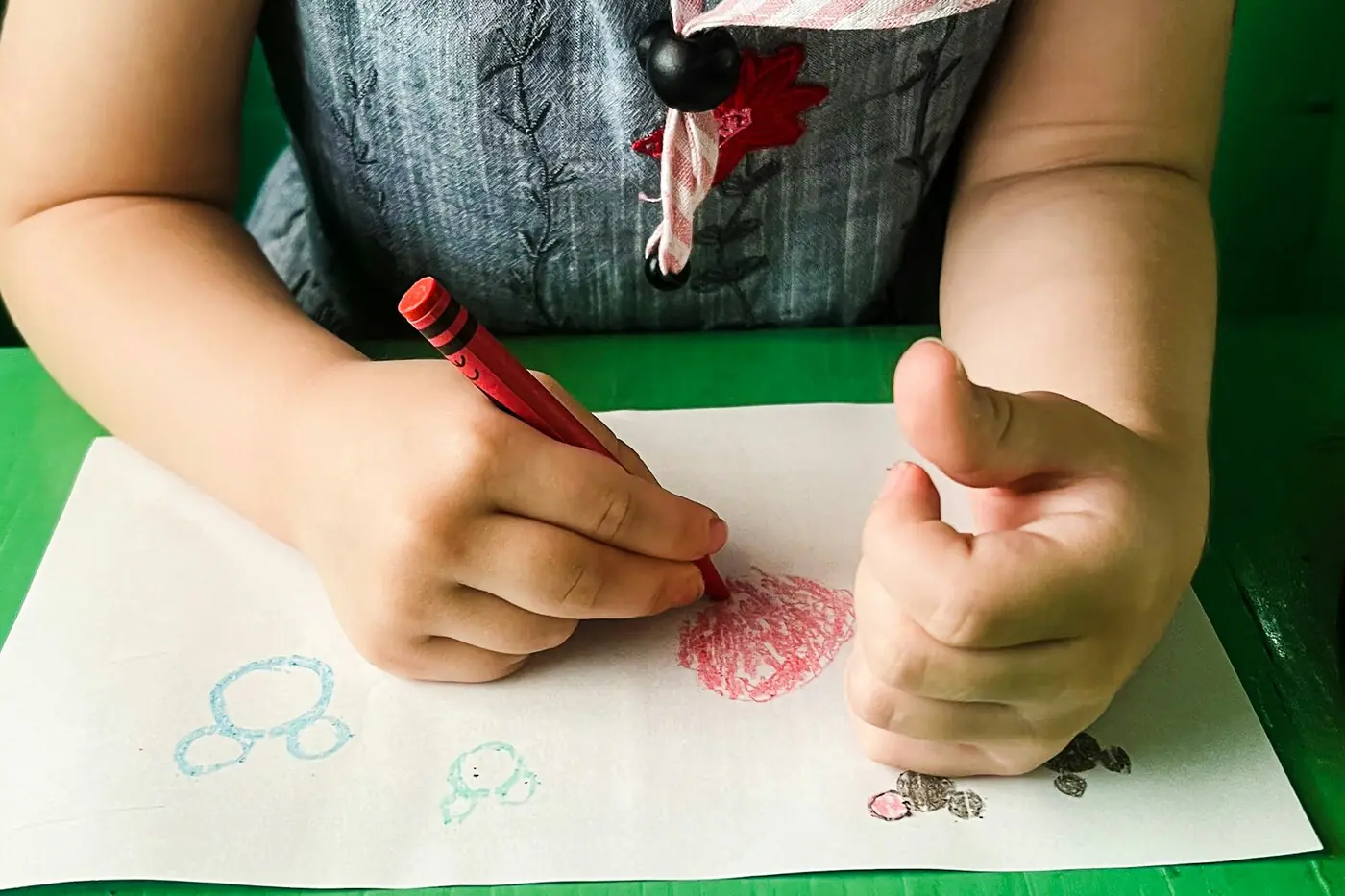 Close overhead view of a small child's hands gripping a red crayon, scribbling on white paper at a green table