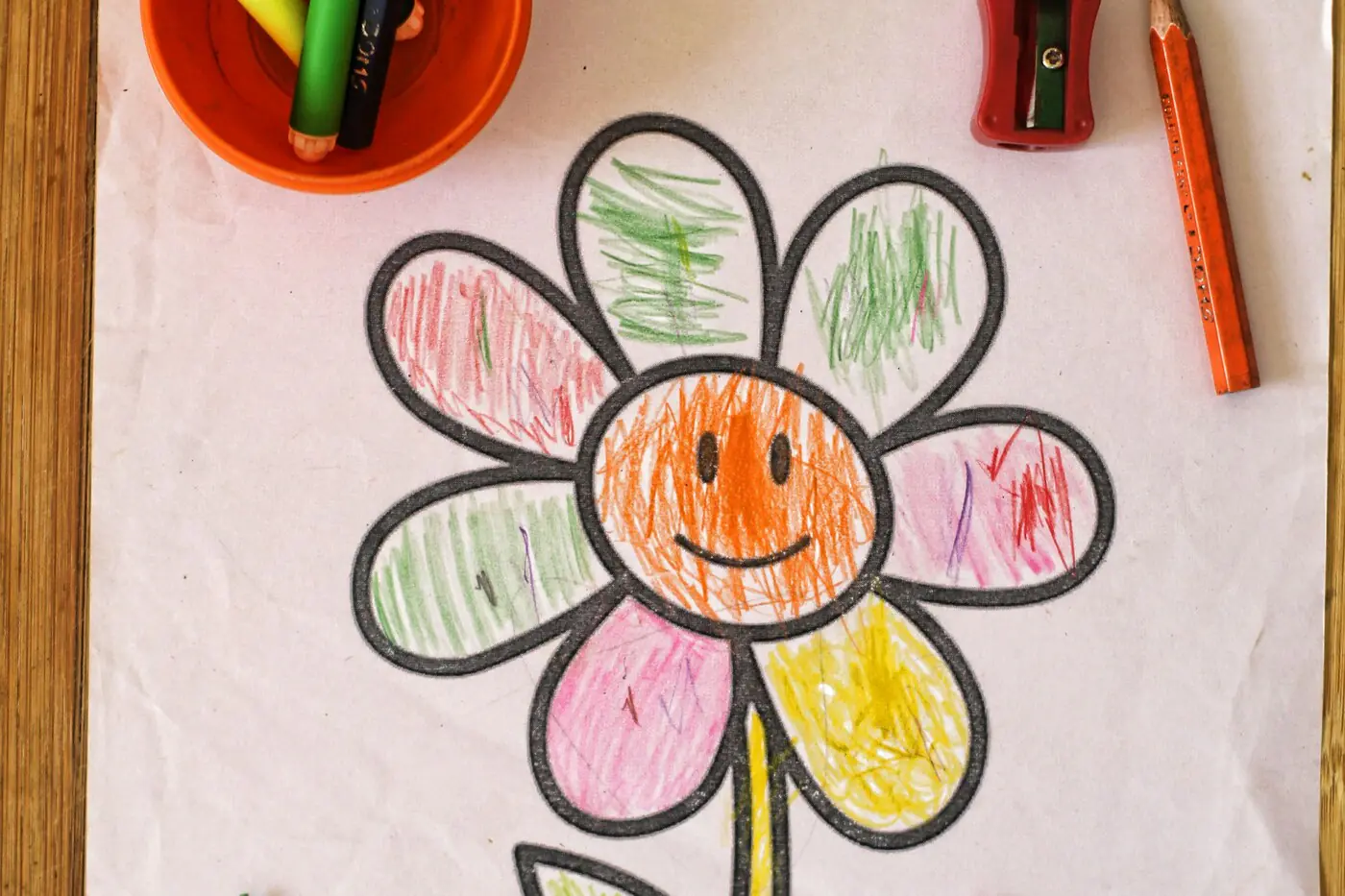A child's crayon drawing of a smiling flower on white paper, lying flat on a wooden table with colored pencils and a sharpener beside it