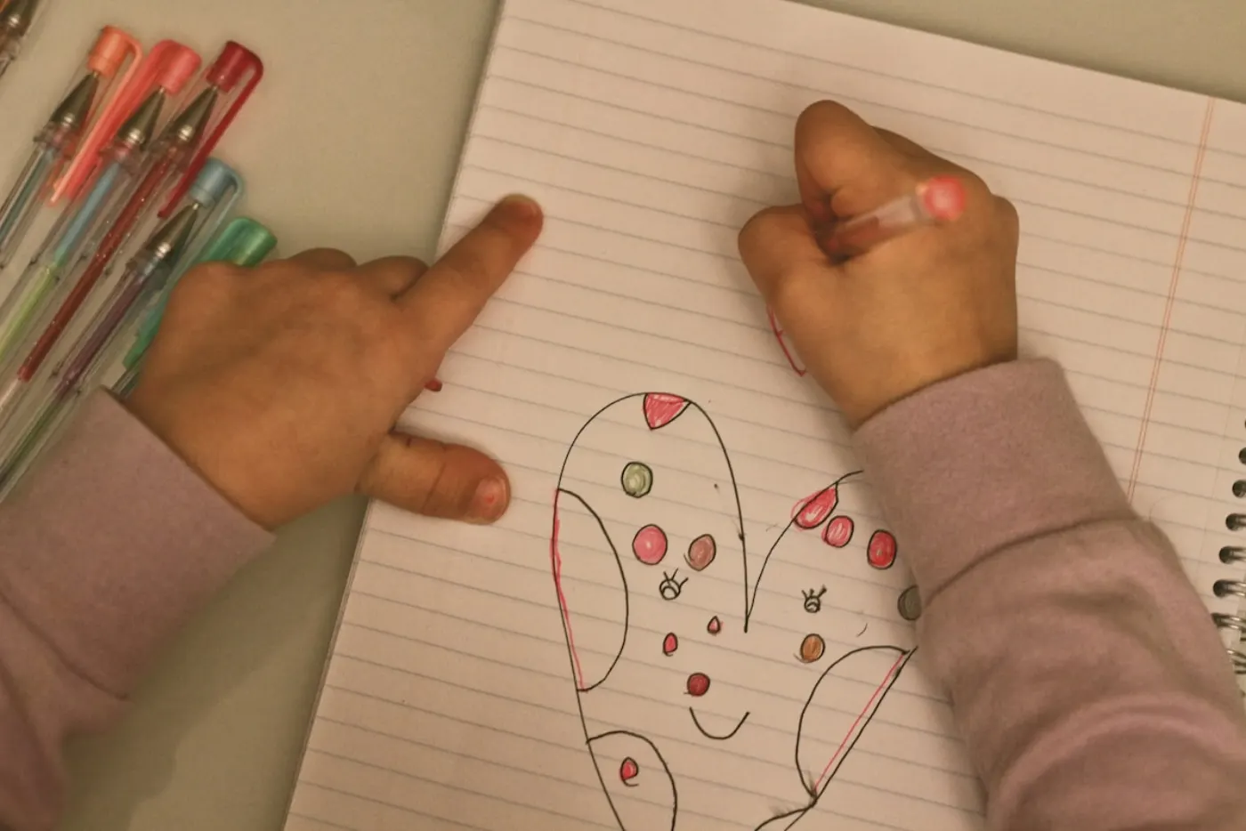 A young child's hands in a pink sweater drawing a heart-shaped creature with a smile in a lined notebook, a row of colored gel pens beside the page
