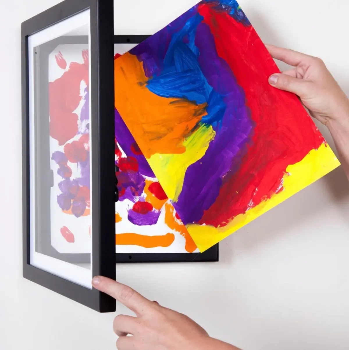 Hands sliding a bright child's painting into a hinged front-opening art frame mounted on a white wall, with another painting already stored behind it