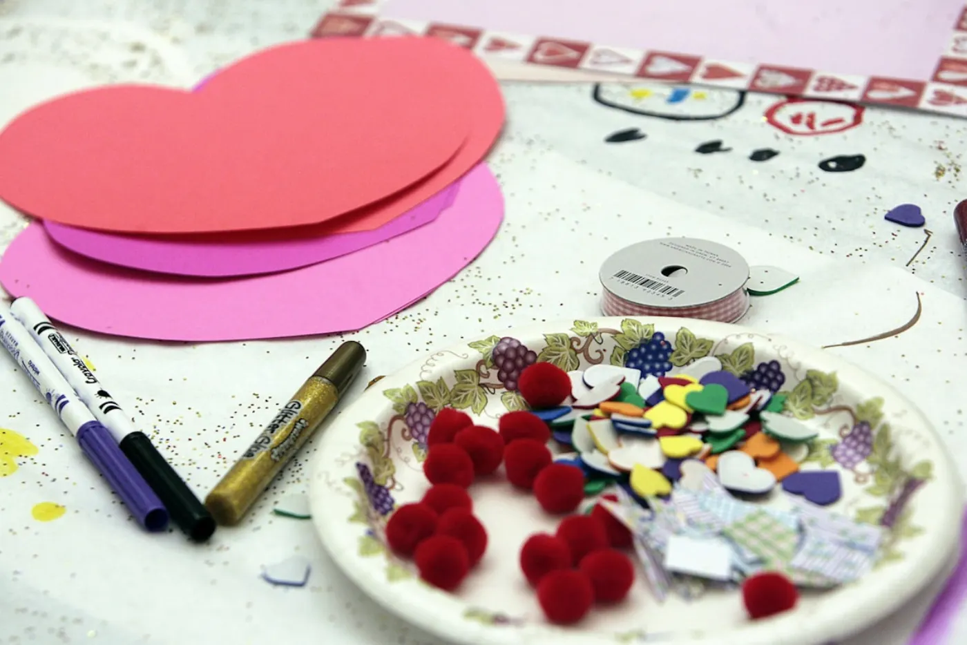 Valentine craft supplies spread on a table: red and pink paper hearts, markers, glitter glue, ribbon, and a paper plate holding red pom poms and foam heart stickers