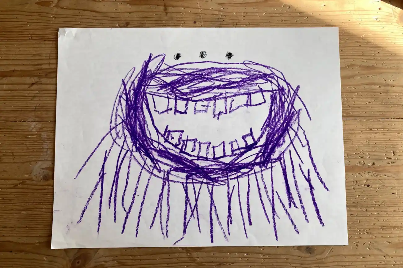 A child's crayon drawing of a purple many-legged monster with three eyes and a wide toothy smile on white paper, alone on a wooden table in warm light