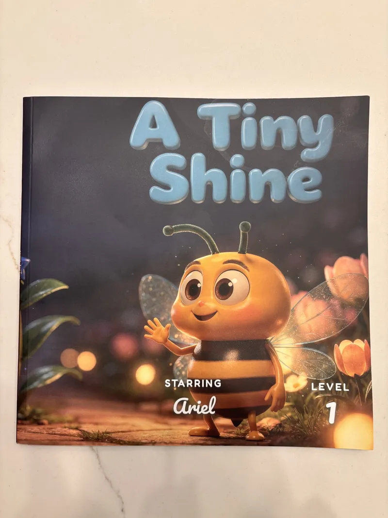 A printed DoodleTale softcover storybook lying on a pale table, titled A Tiny Shine, with a bee character on the cover and the words starring Ariel underneath