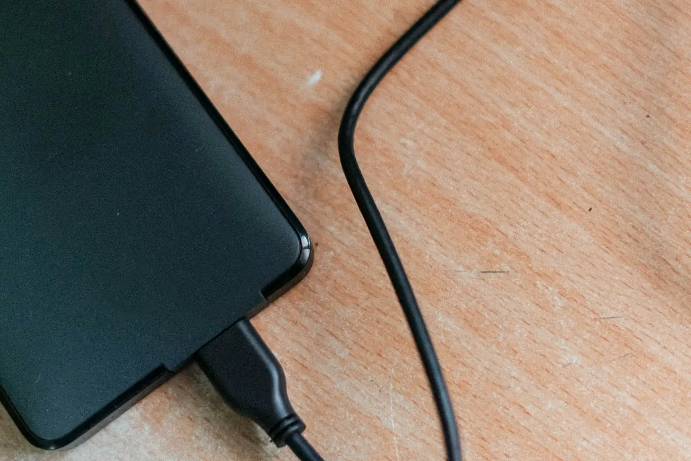 A plain unbranded black portable hard drive lying on a light wood desk with its USB cable plugged in and curving away across the grain