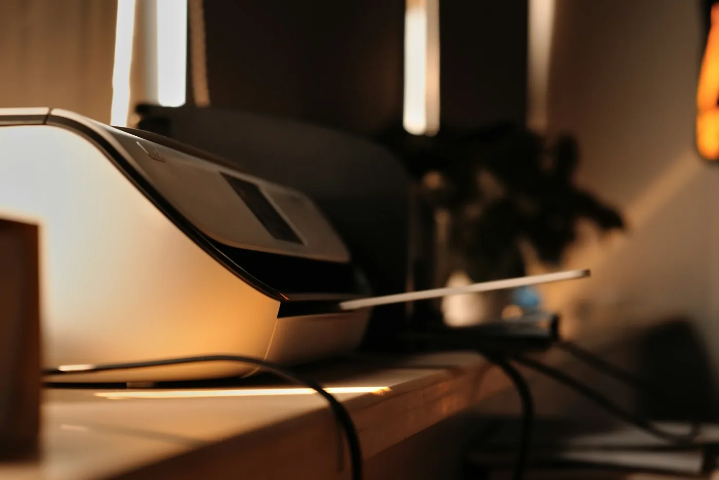 A home flatbed scanner and printer on a wooden desk in low golden window light, its lid raised, a cable trailing off the desk edge